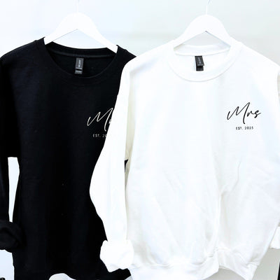 A pair of soft, personalized wedding sweatshirts, one black and one white, each featuring custom script lettering on the chest, hung on white hangers.
