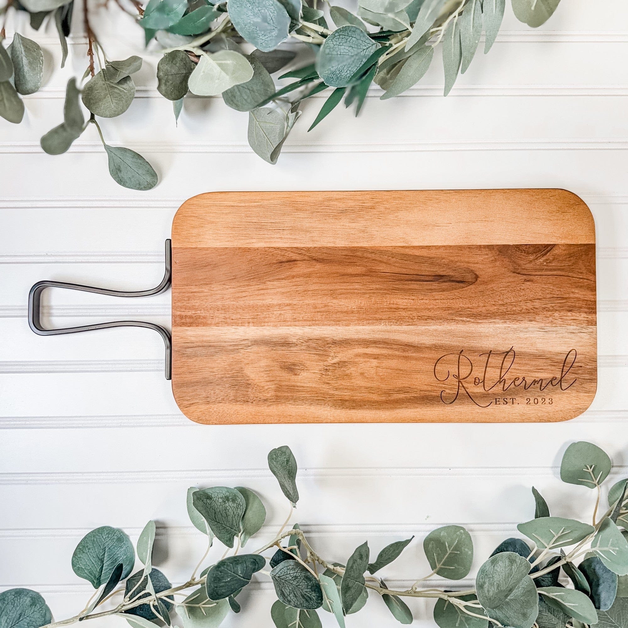 A rectangular natural wood serving board with rounded corners and a dark metal handle, featuring custom laser engraving, displayed as a personalized wedding or anniversary gift.