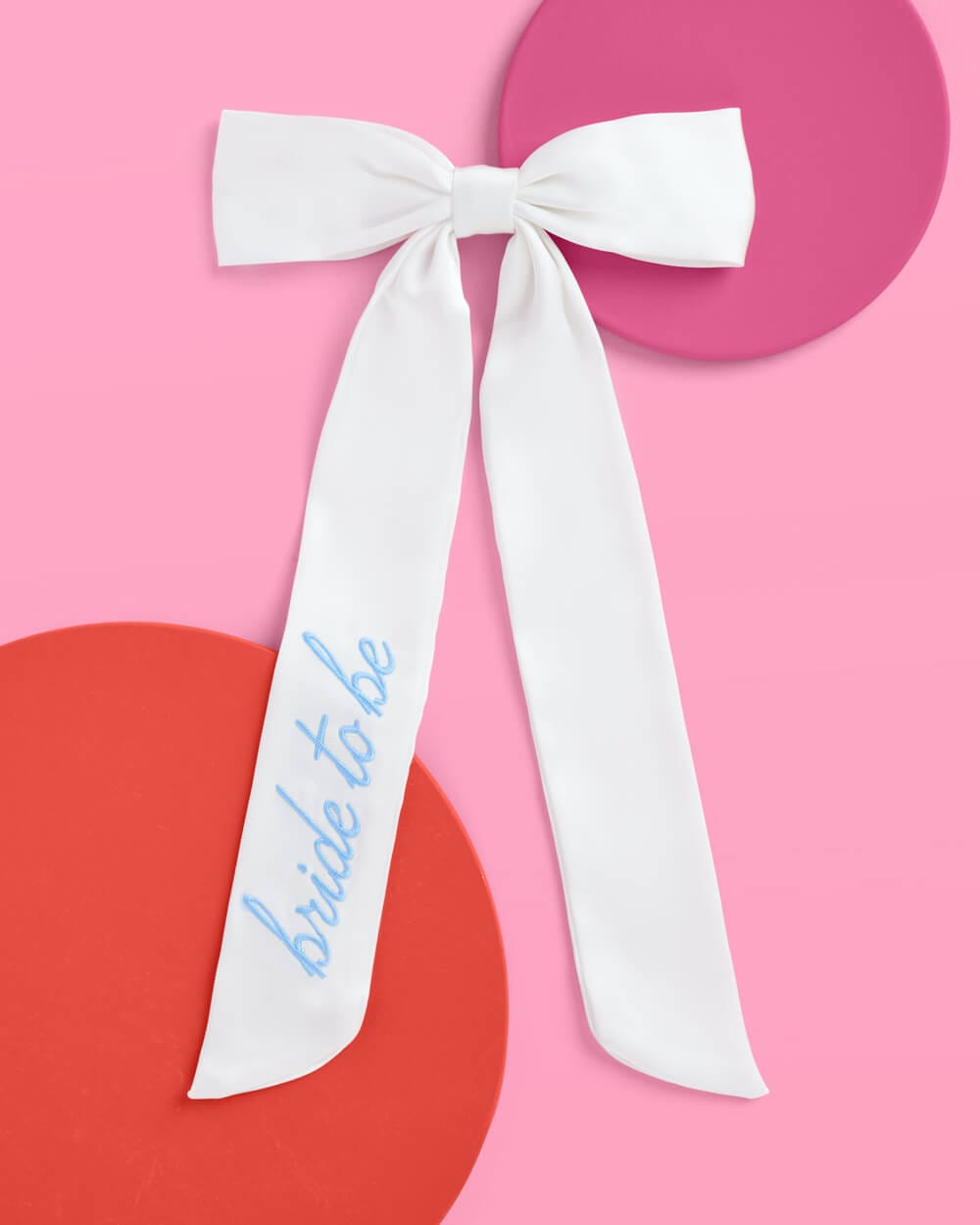 White satin bow with blue "Something Blue" embroidery on trailing ribbon against pink background