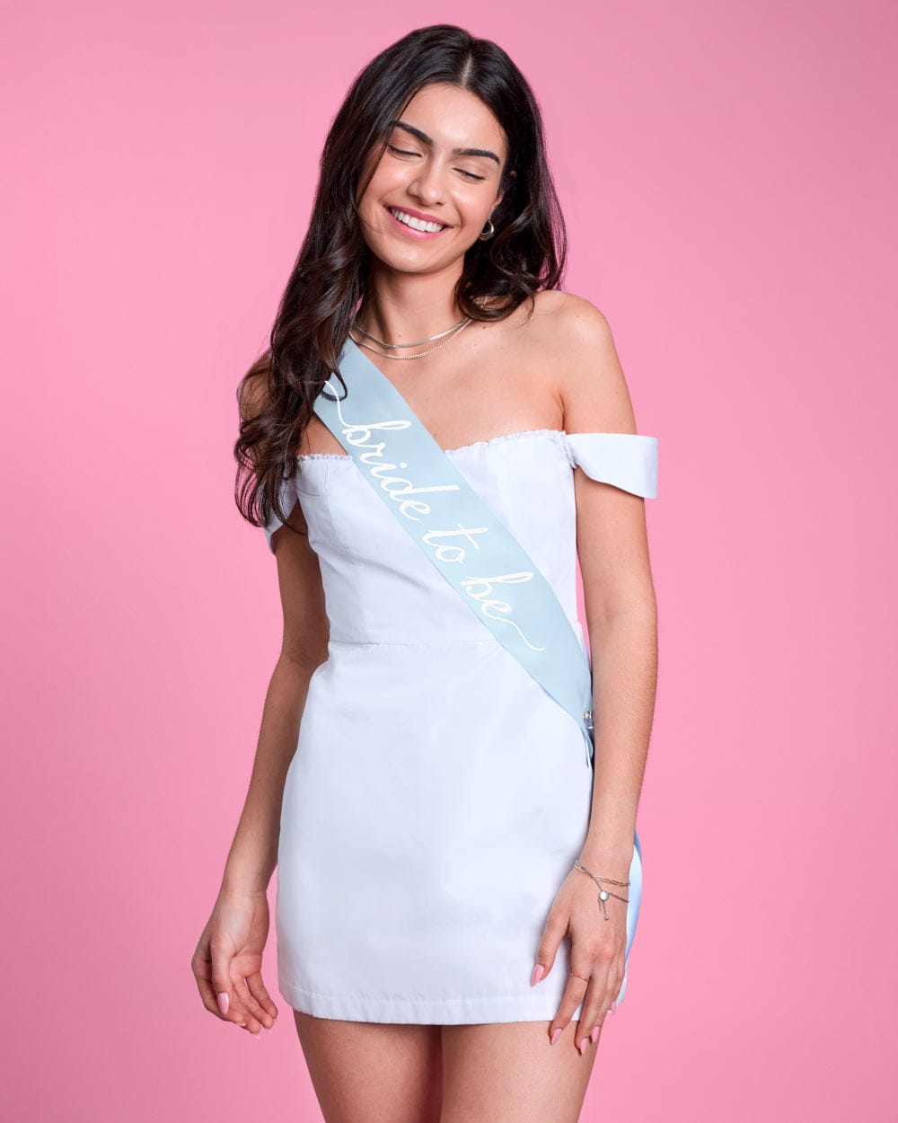 Woman wearing white off-shoulder dress with light blue satin sash across chest against pink background