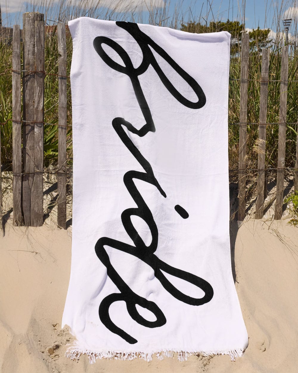 White cotton beach towel with black script text reading "bride" hung on sandy beach fence.