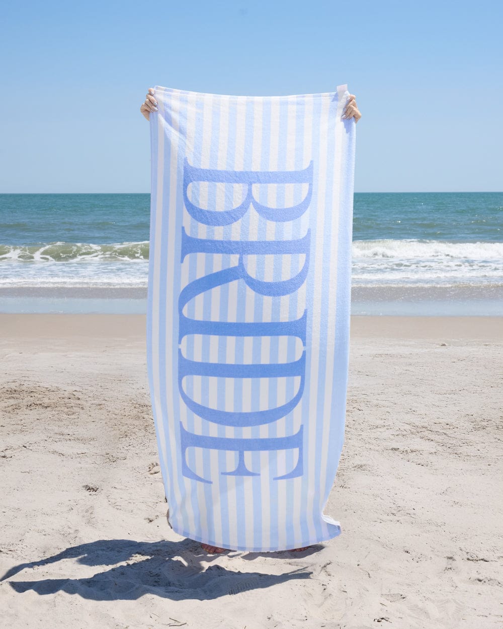 Blue and white striped beach towel with "BRIDE" text held up on sandy beach with ocean waves in background