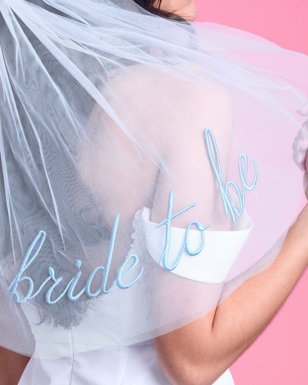 White tulle veil with blue embroidered "bride to be" text, worn over white cup against pink background.