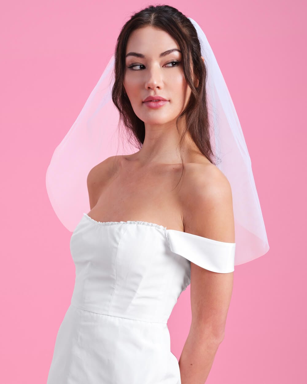 Woman wearing white off-shoulder wedding dress with sheer tulle veil against pink background