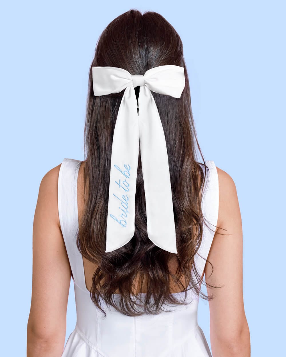 White silk hair bow with personalized "something blue" text, worn in long dark hair against blue background.