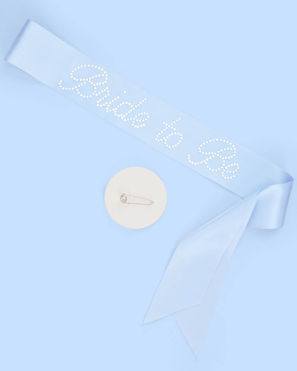 Light blue bridal sash with rhinestone text and matching cream circular pin with safety pin design on blue background.