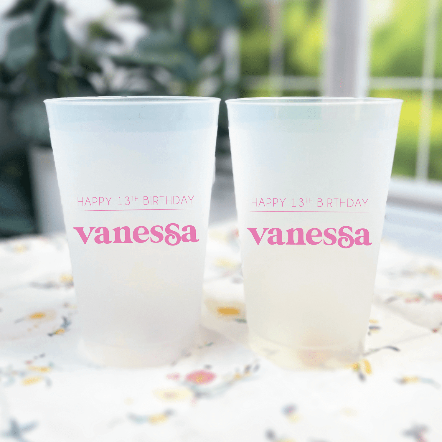 Two frosted plastic cups with pink text reading "Happy 13th Birthday Vanessa" displayed on outdoor table.