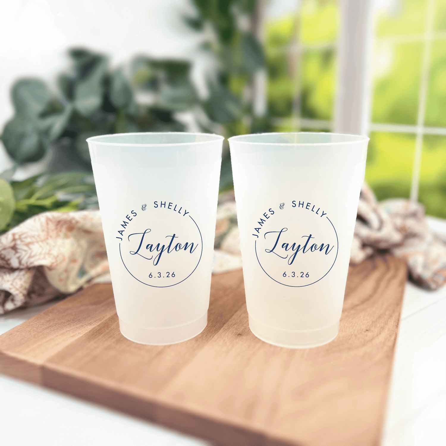 Two frosted cups with "His & Hers Shelby Taylow 4.3.24" printed in circular monogram design, displayed on wooden board with greenery.