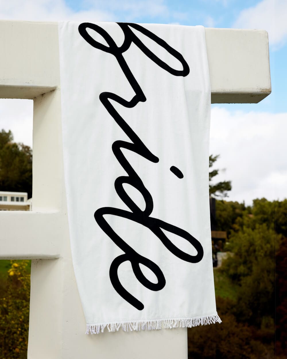 White cotton beach towel with black script "bride" text and fringe trim, draped on railing.