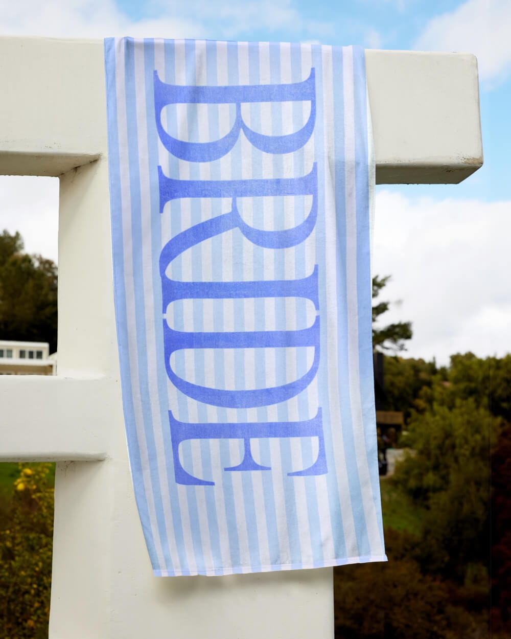 Periwinkle and white striped beach towel with large "BRIDE" text in blue, hanging outdoors.