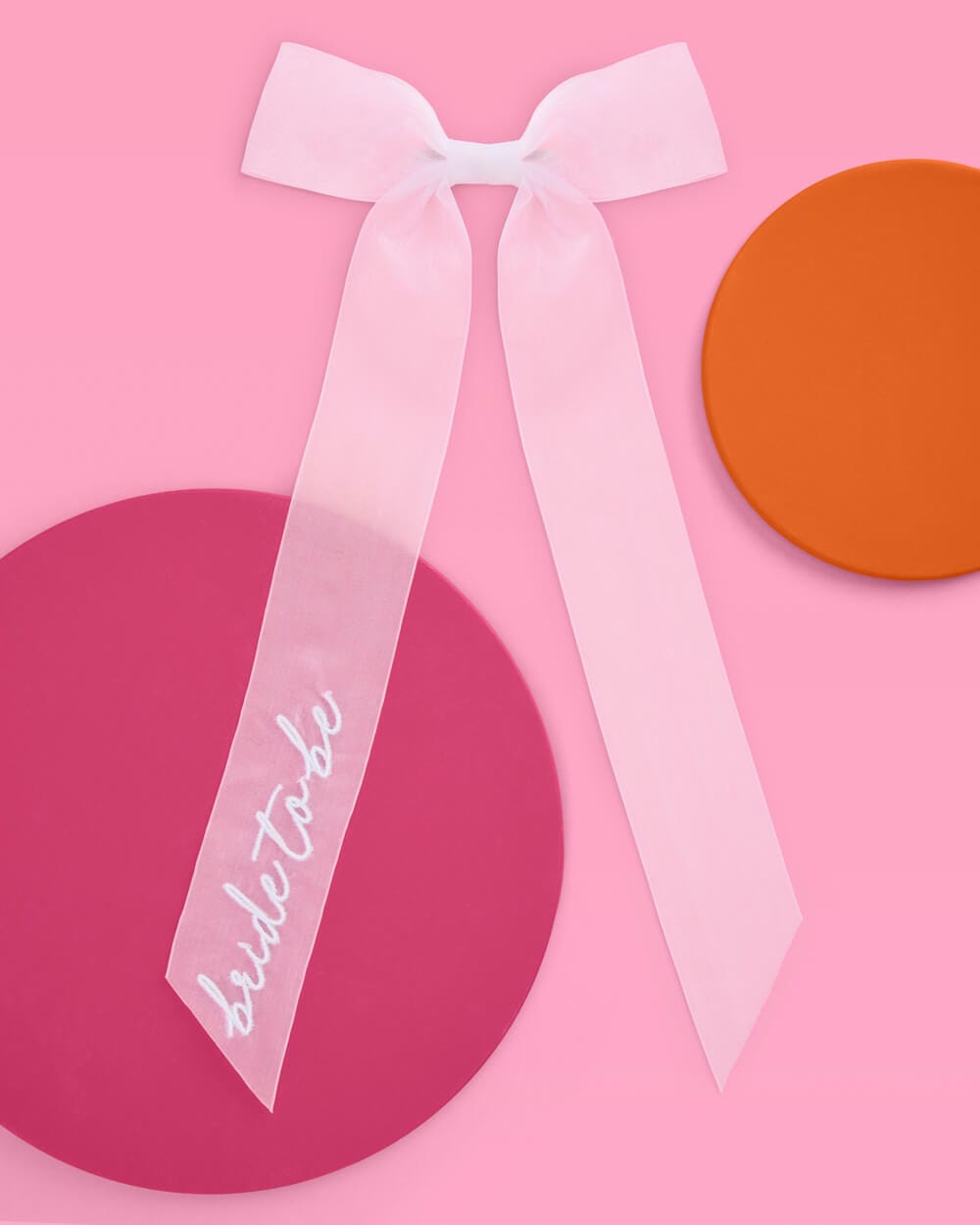 Pink satin bow with white "bride to be" embroidered script on left streamer against pink background with decorative circles.