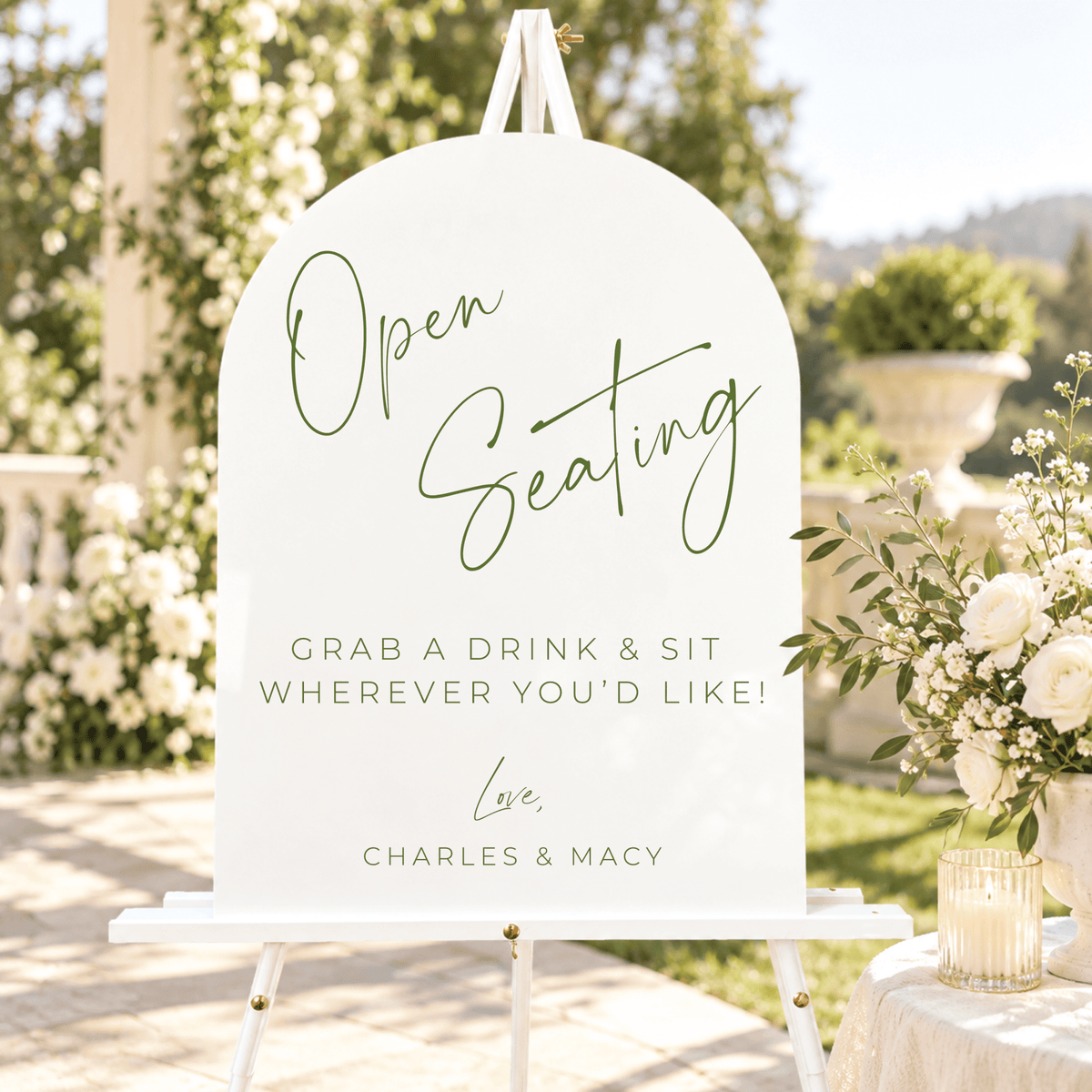 Arched open seating wedding sign with green script lettering, displayed outdoors on a white easel beside elegant wedding florals.