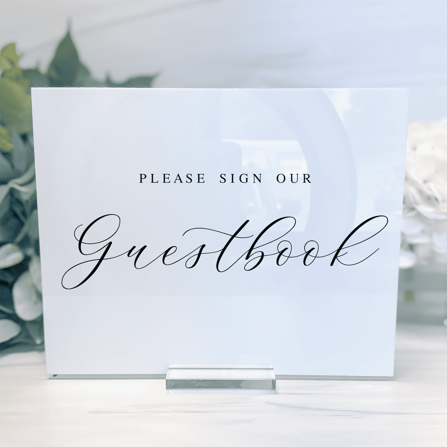White acrylic guestbook sign with "Please Sign Our Guestbook" text in elegant script, displayed on a clear stand with greenery backdrop.