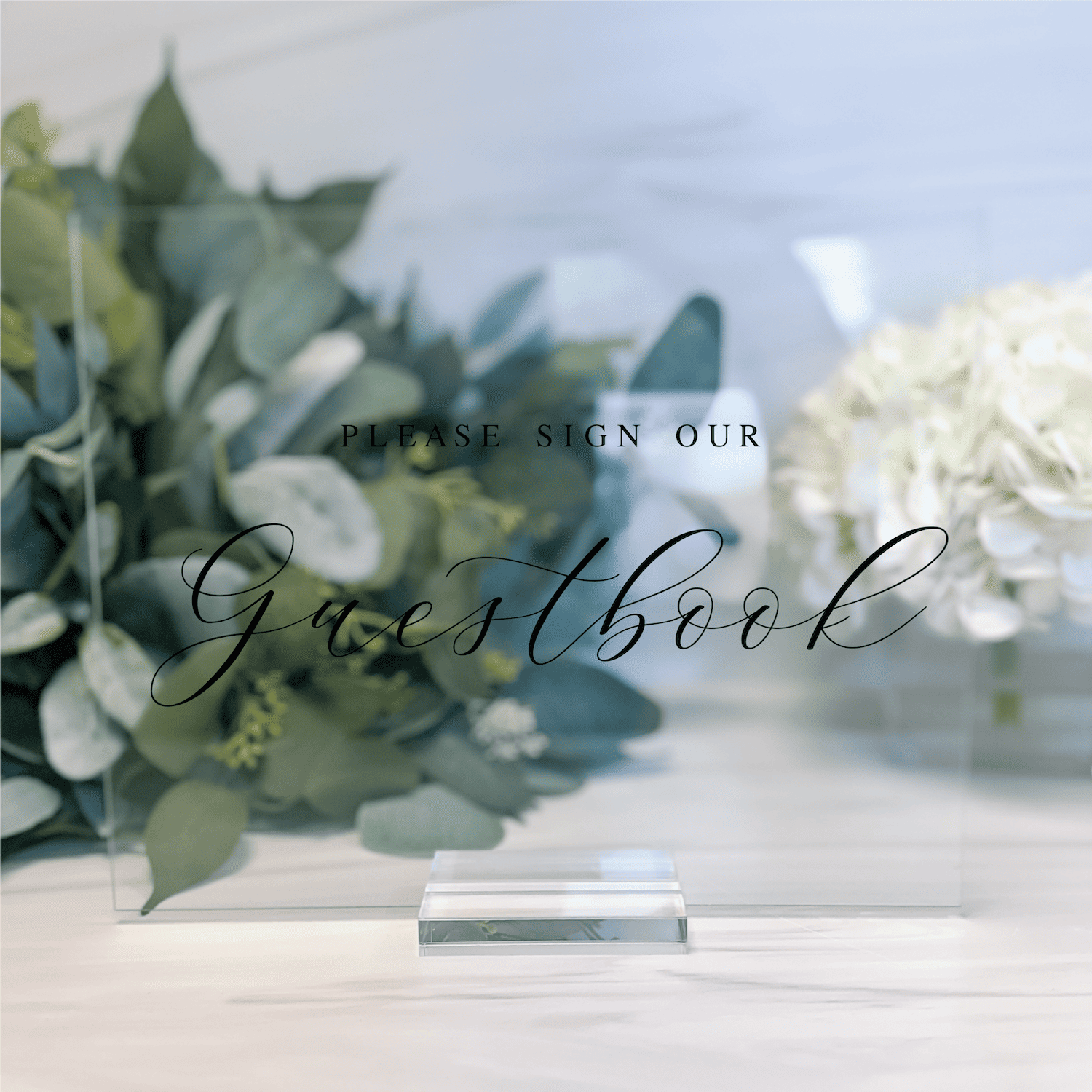 Frosted acrylic guestbook sign with "Please Sign Our Guestbook" text, displayed on table with greenery and white flowers.