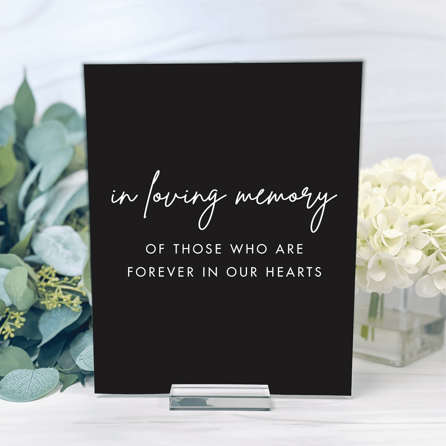 Black acrylic table sign with white script text reading "in loving memory of those who are forever in our hearts" displayed between eucalyptus and white flowers.
