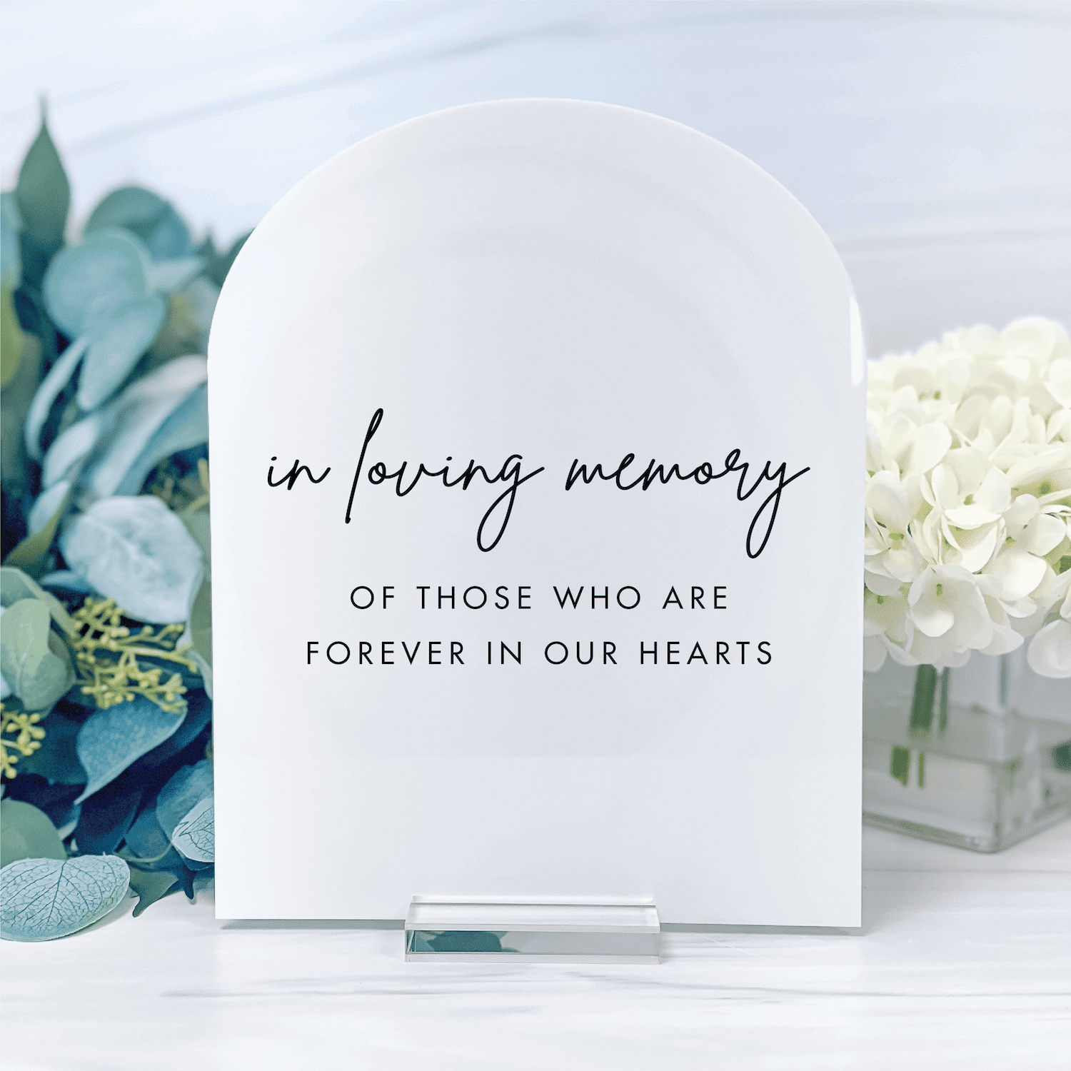 White arched acrylic table sign with "in loving memory of those who are forever in our hearts" text, displayed with greenery and white flowers.