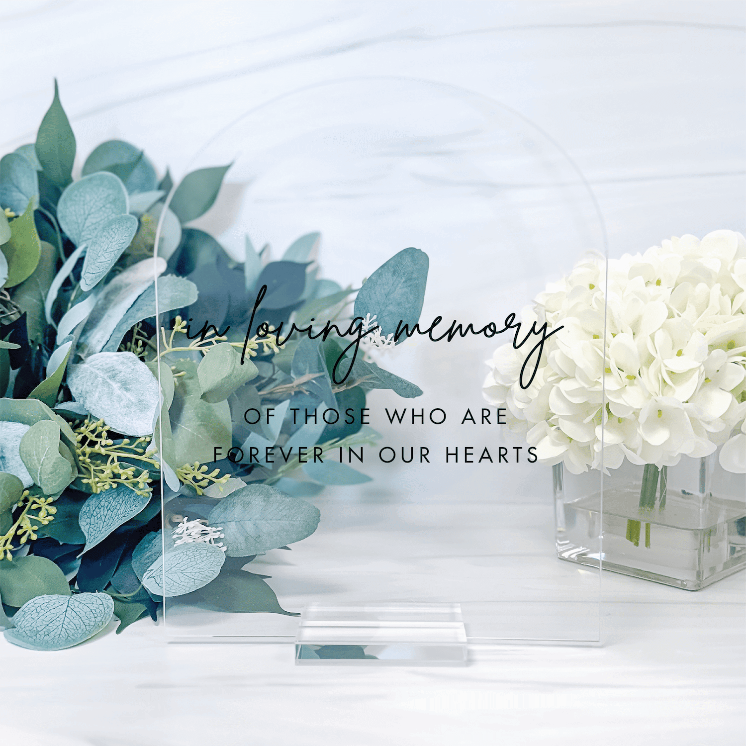 Clear acrylic table sign with "in loving memory" script text and "of those who are forever in our hearts" message, displayed among white flowers and eucalyptus greenery.