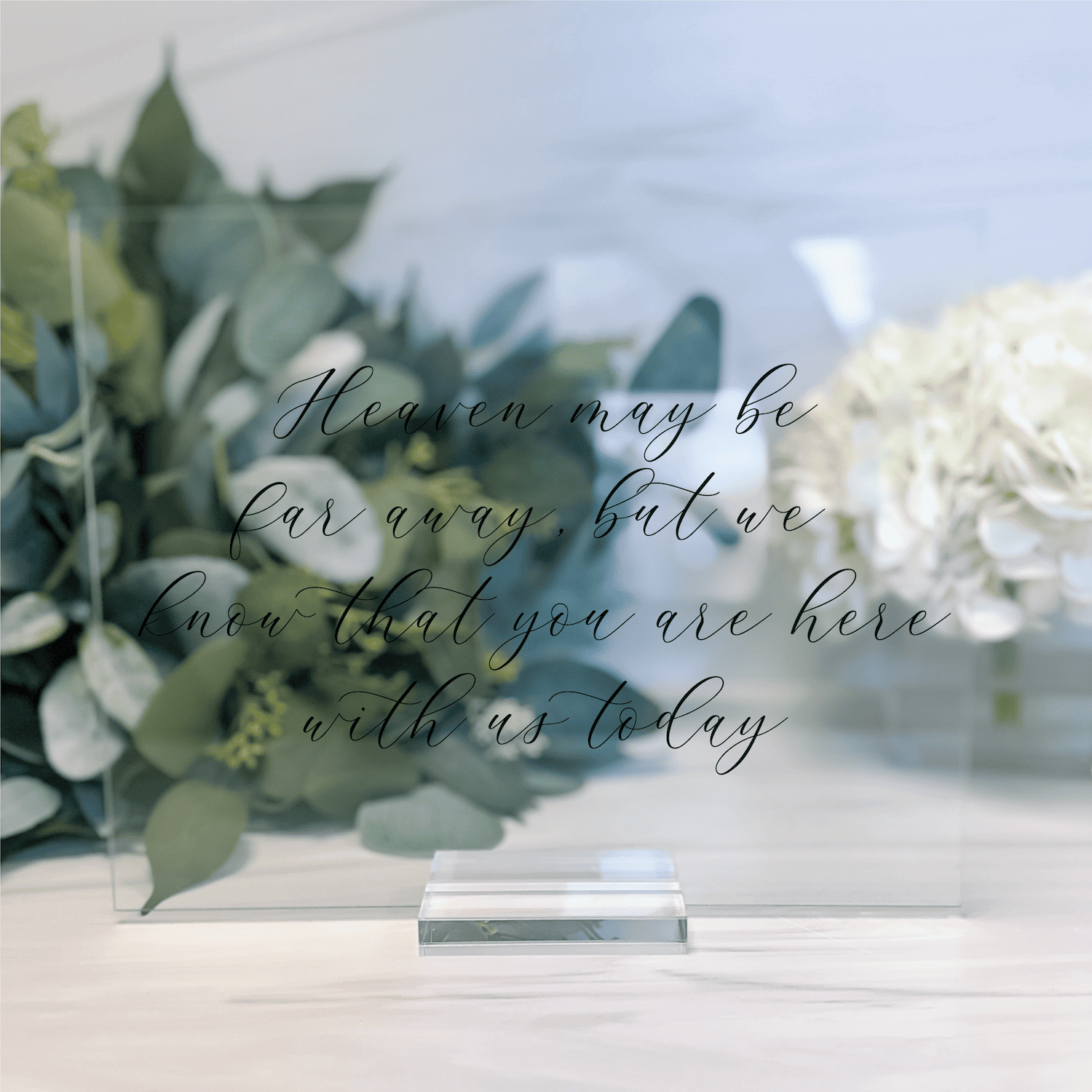 Clear acrylic wedding sign with script text reading "Heaven may be forever but you are here with us today" displayed with white flowers and greenery.
