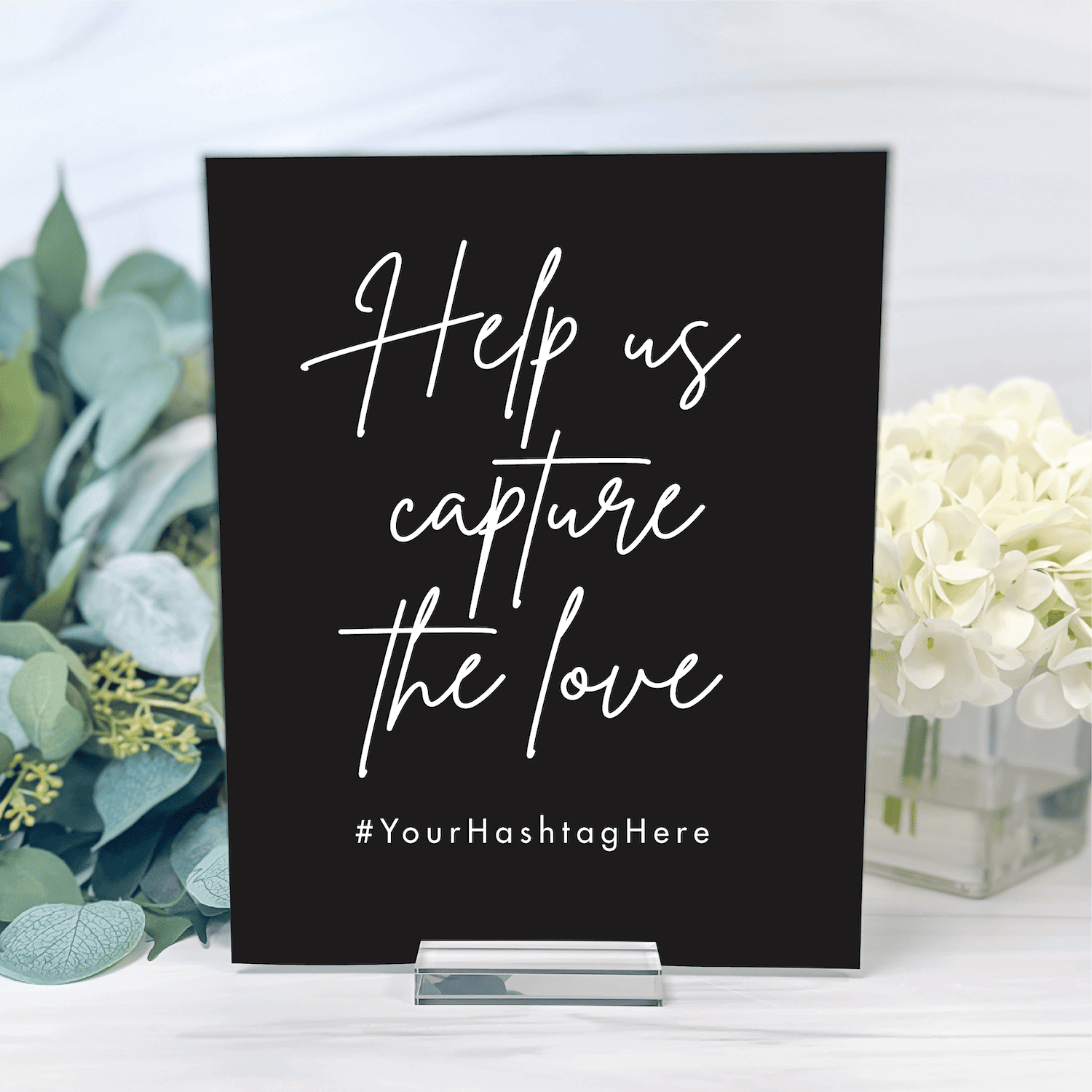 Black acrylic sign with white handwritten text reading "Help us capture the love" and "#YourHashtagHere" surrounded by eucalyptus and white flowers.