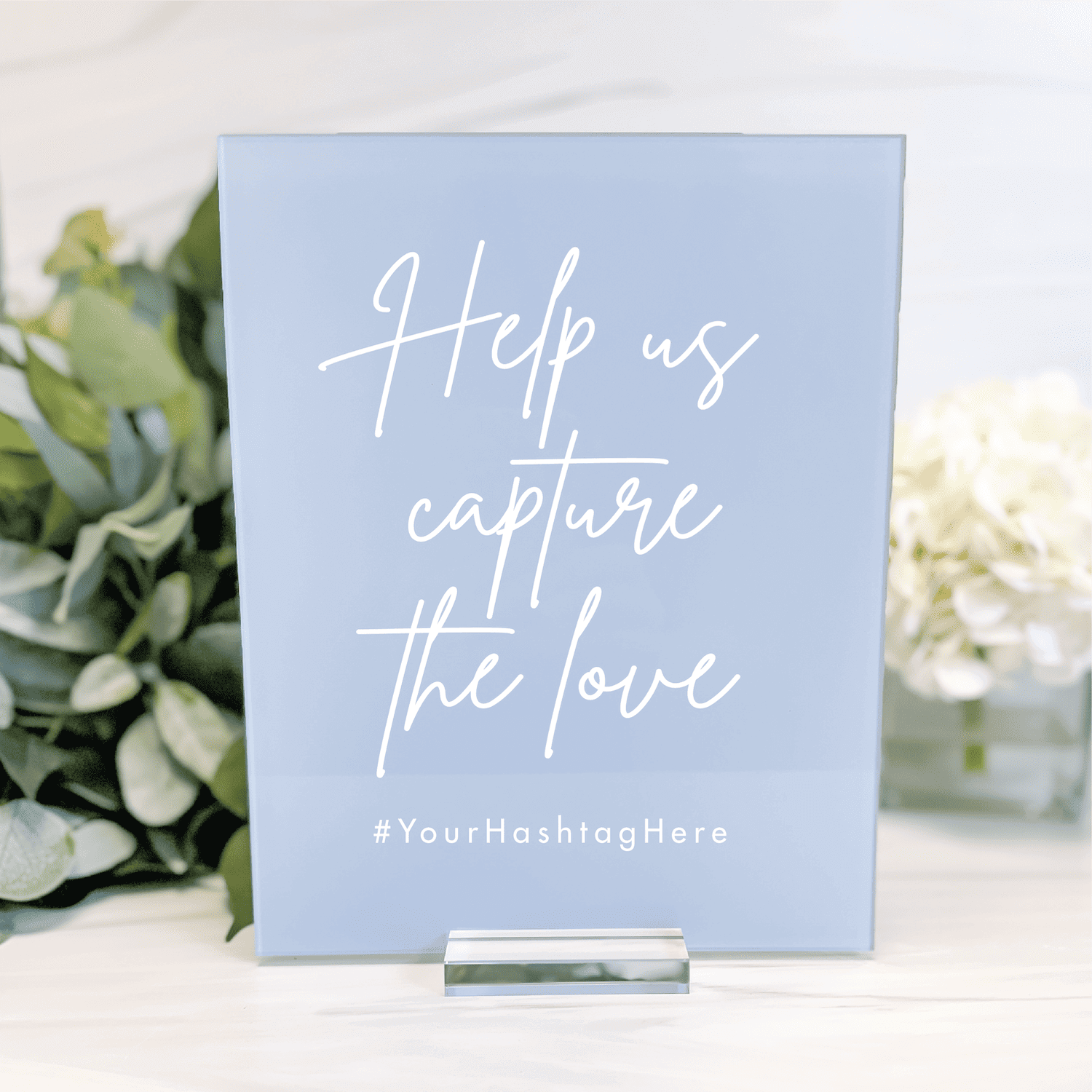 Light blue acrylic wedding sign with white script text reading "Help us capture the love" and hashtag placeholder, displayed with flowers