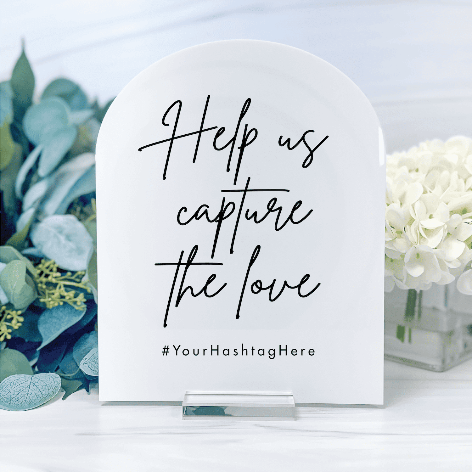 White acrylic wedding sign with handwritten-style text reading "Help us capture the love" and customizable hashtag space, surrounded by eucalyptus and white flowers.