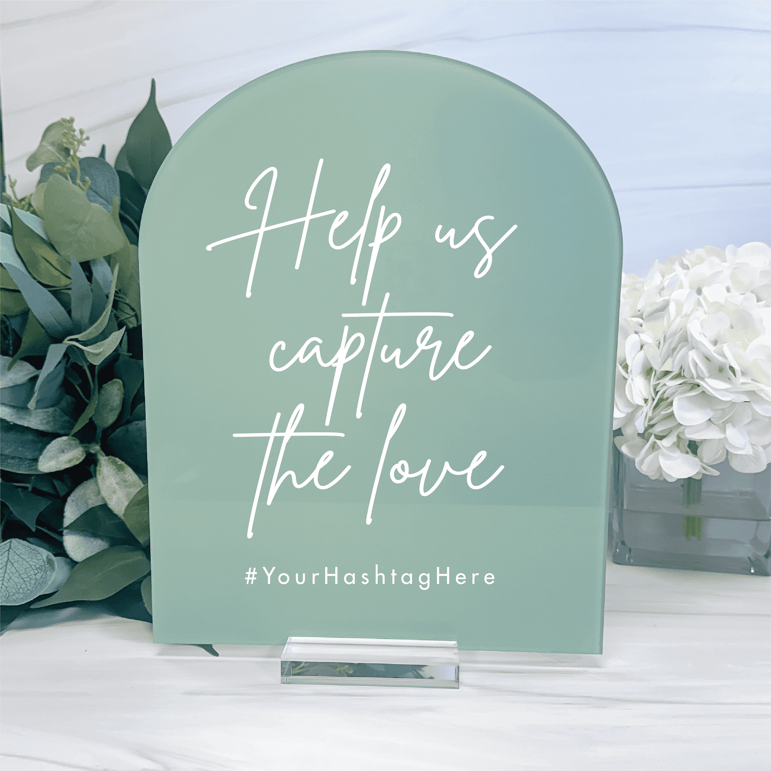 Sage green arch-shaped acrylic sign with white script text reading "Help us capture the love" and hashtag placeholder, displayed with greenery and white flowers.