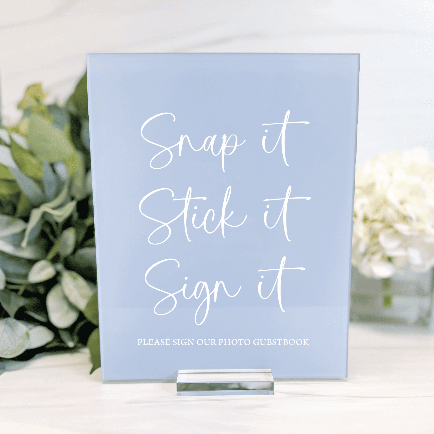 Light blue acrylic sign with white cursive text reading "Snap it Stick it Sign it" and smaller text below for photo guestbook instructions, displayed on a stand with flowers.