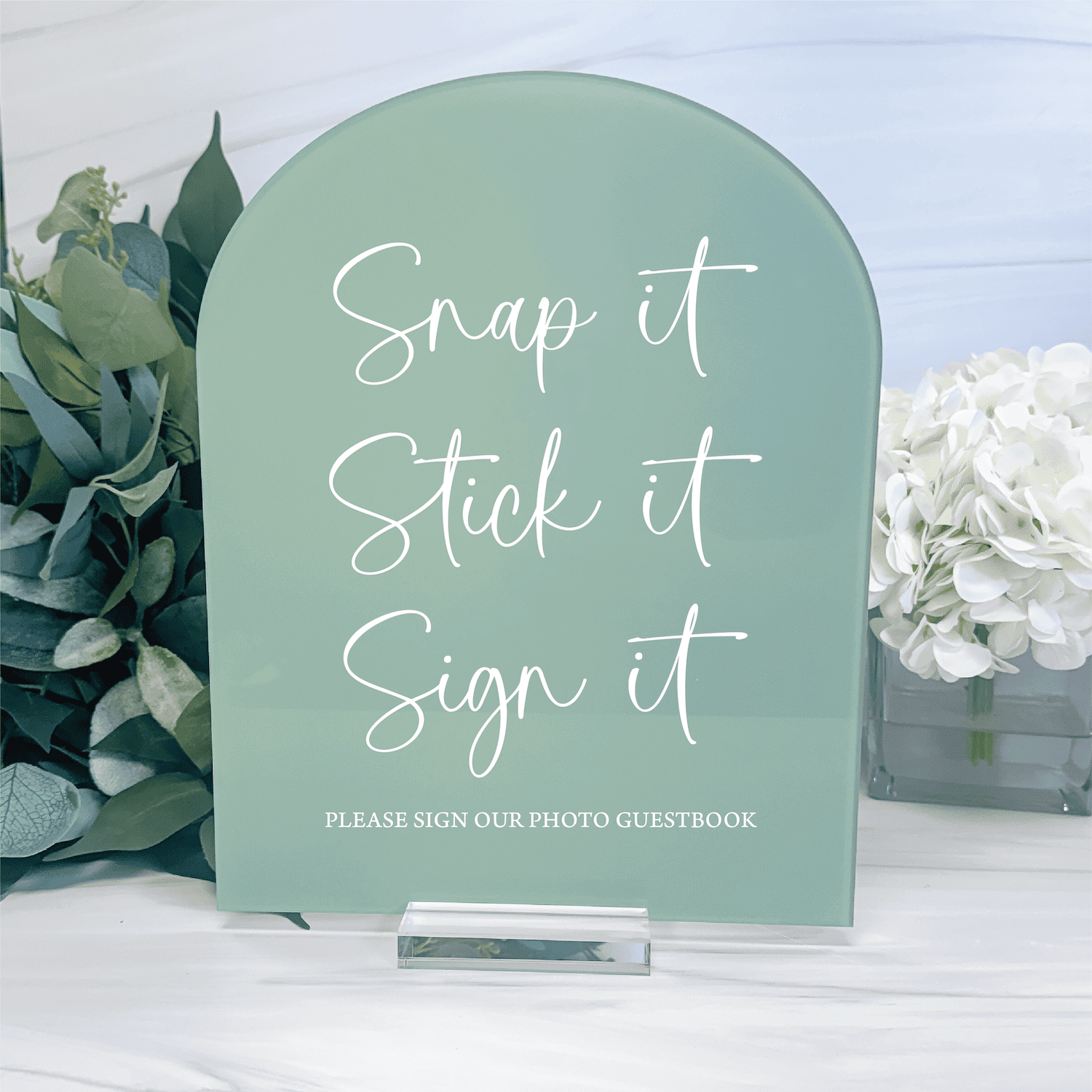 Sage green arch-shaped acrylic sign with white script text reading "Snap it Stick it Sign it" and "Please sign our photo guestbook"