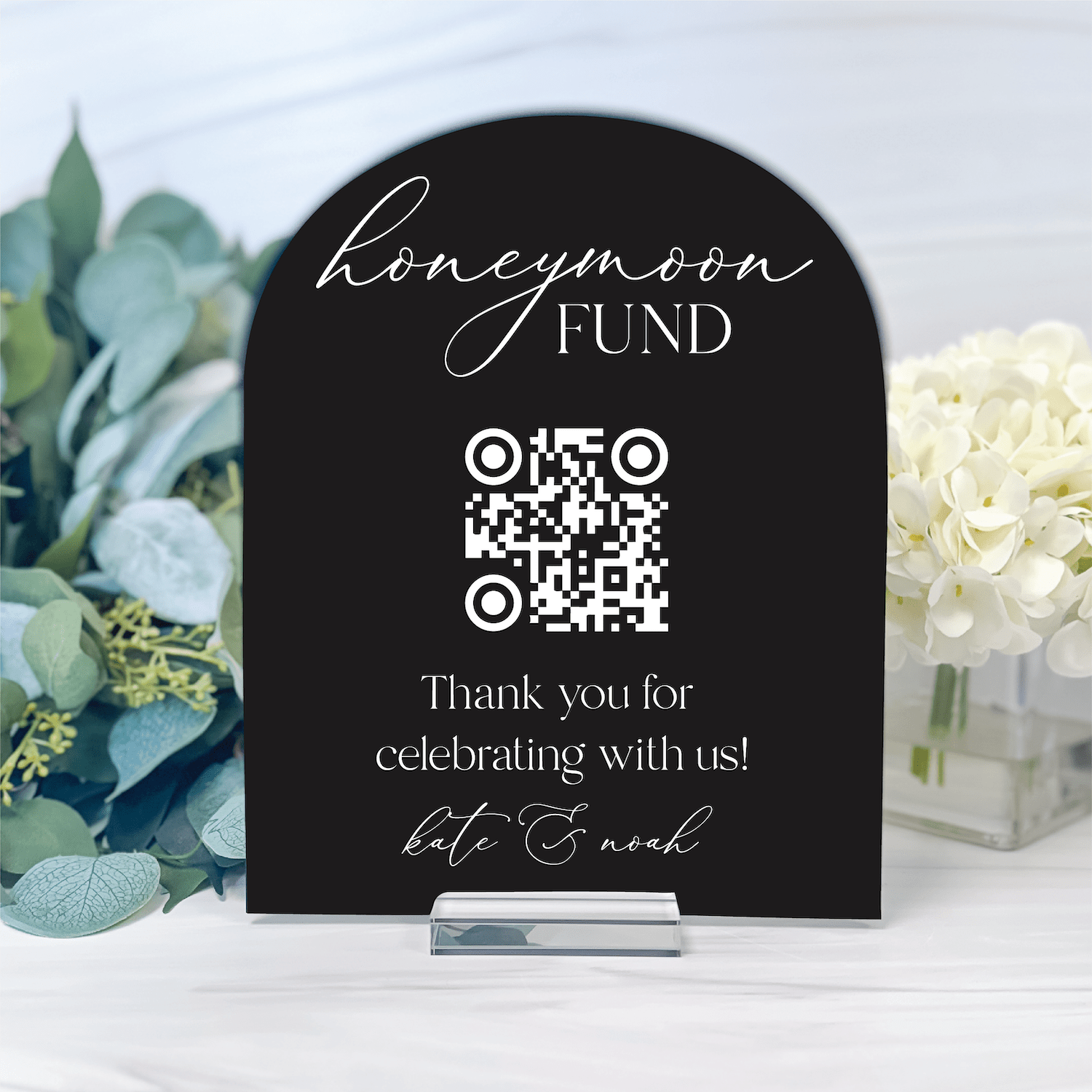 Black arched wedding sign with "Honeymoon Fund" text, QR code, and personalized names, displayed with flowers and greenery