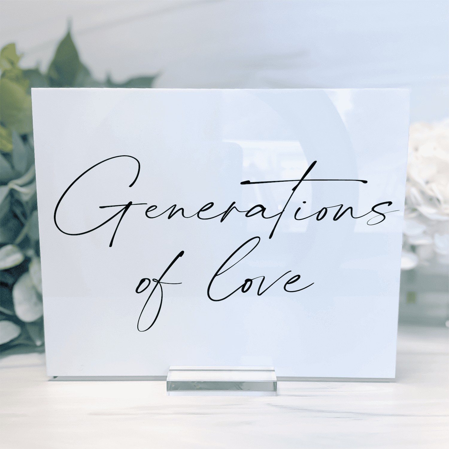 White acrylic sign with "Generations of Love" in elegant script lettering, displayed on clear stand with greenery background.
