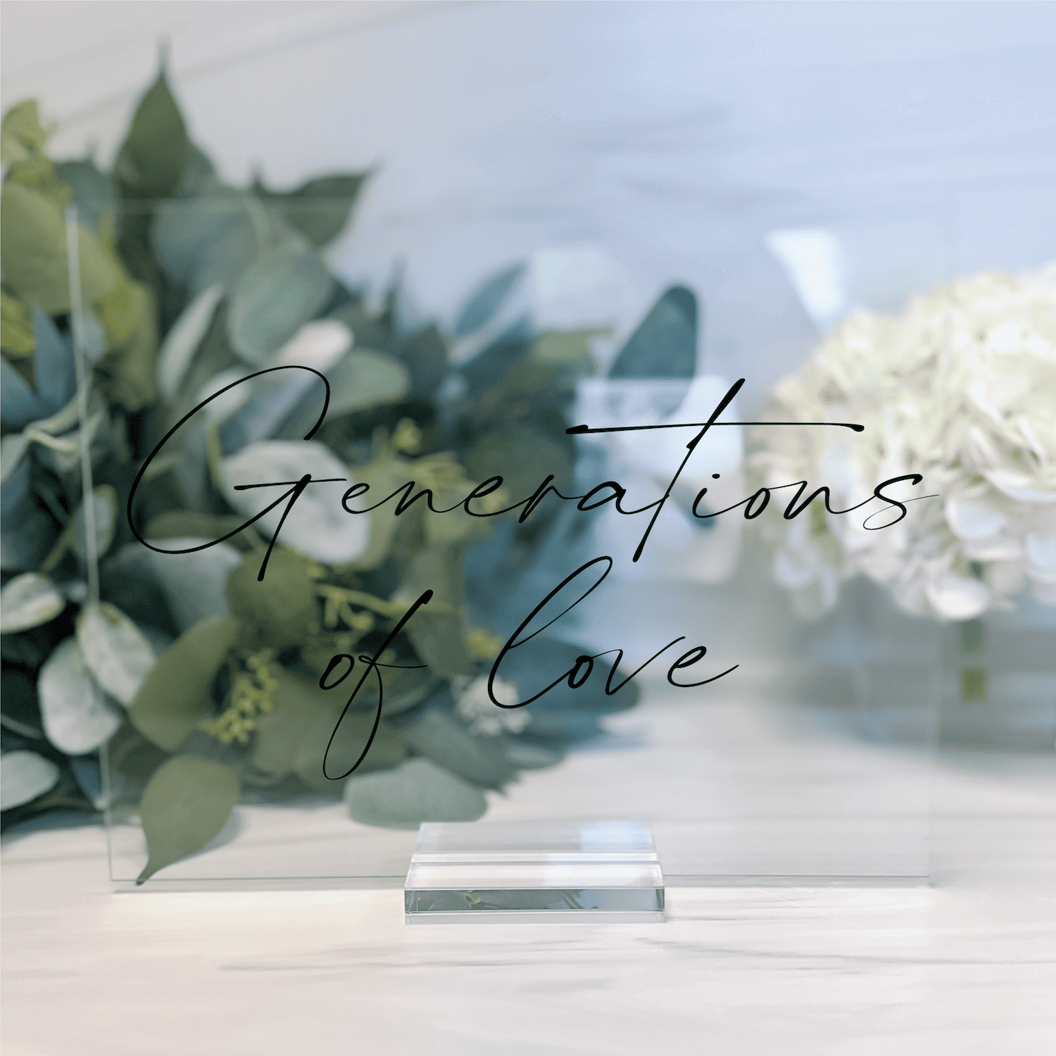 Frosted acrylic wedding sign with "Generations of Love" script text, displayed on table with green eucalyptus and white flowers.
