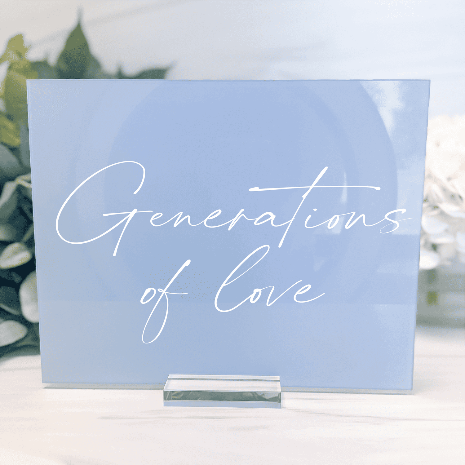 Light blue acrylic sign with white script text reading "Generations of Love" on a silver stand, displayed outdoors with greenery in background.