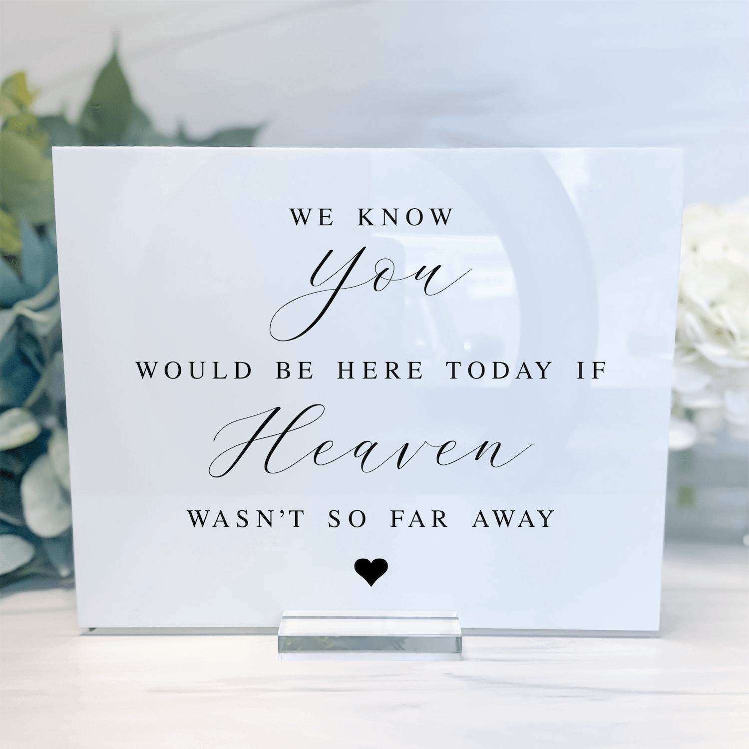 White acrylic memorial sign with black script and serif text reading "We know you would be here today if Heaven wasn't so far away" with small heart, displayed on stand with floral background.