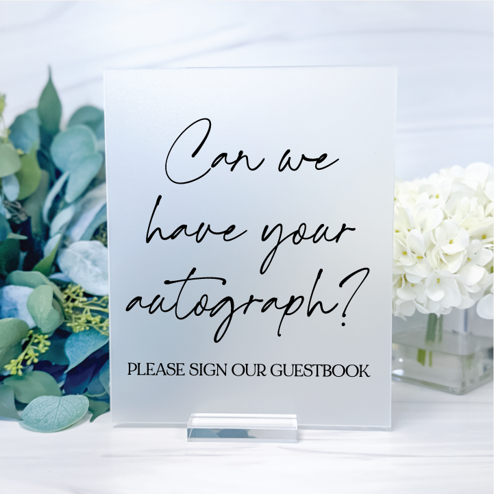 Frosted acrylic wedding guest book sign with black script reading Can We Have Your Autograph, displayed with greenery and white flowers.