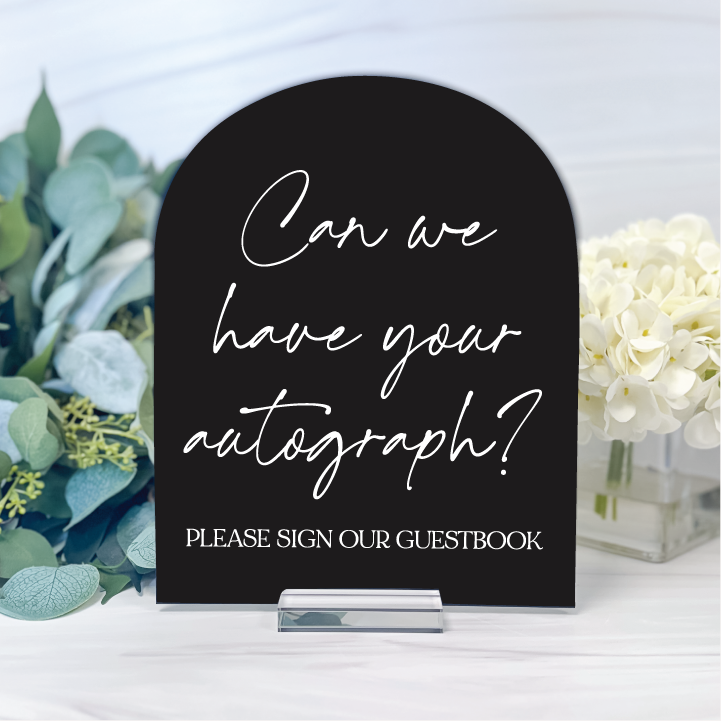 Black arched acrylic wedding guestbook sign with elegant white script reading Can We Have Your Autograph and Please Sign Our Guestbook.