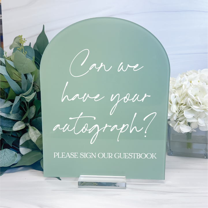 Sage green arched acrylic wedding guest book sign with white script lettering, displayed on a clear stand beside eucalyptus and white flowers.
