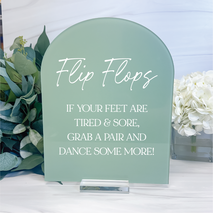 Sage green arched acrylic wedding dancing shoes sign featuring white lettering and a flip flop poem, styled with greenery and white flowers.
