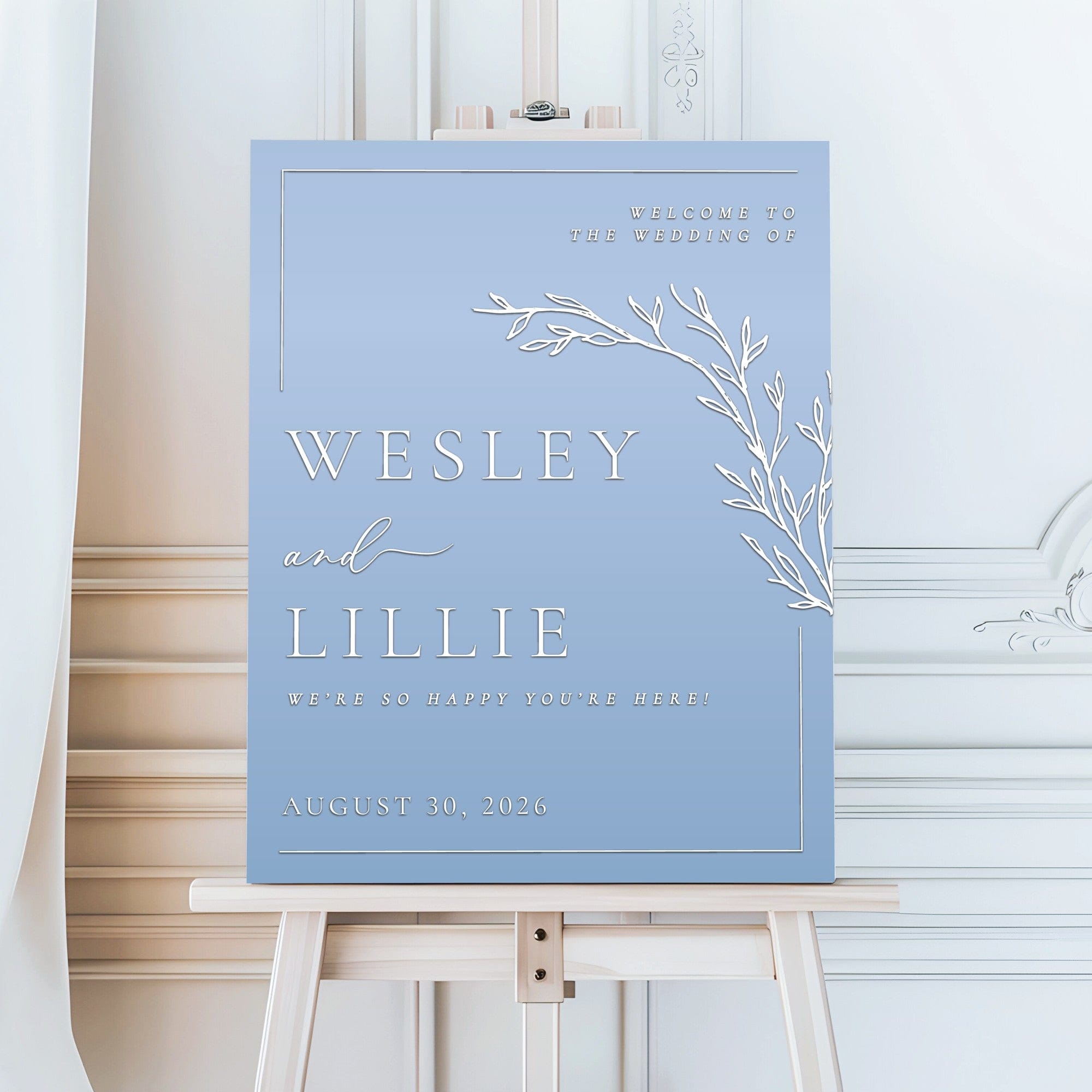 A rectangular wedding welcome sign with a light blue background, featuring a white thin border, a white botanical branch illustration, and white text welcoming guests to the event.