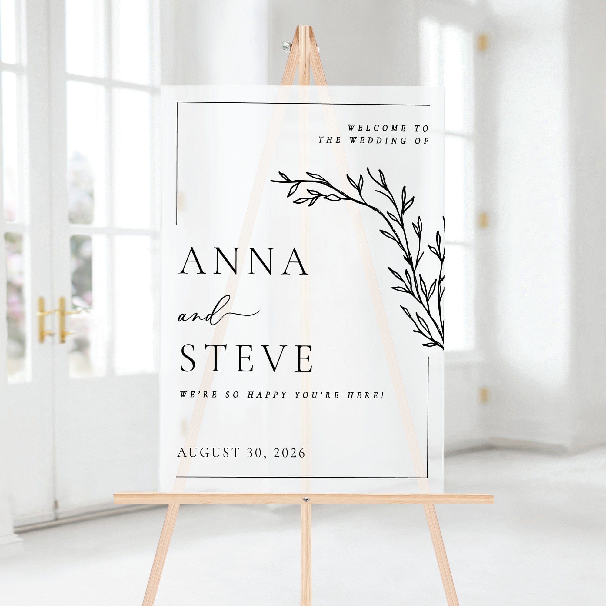 A personalized white acrylic wedding welcome sign featuring elegant black text and a minimalist botanical leaf design, displayed on a wooden easel.