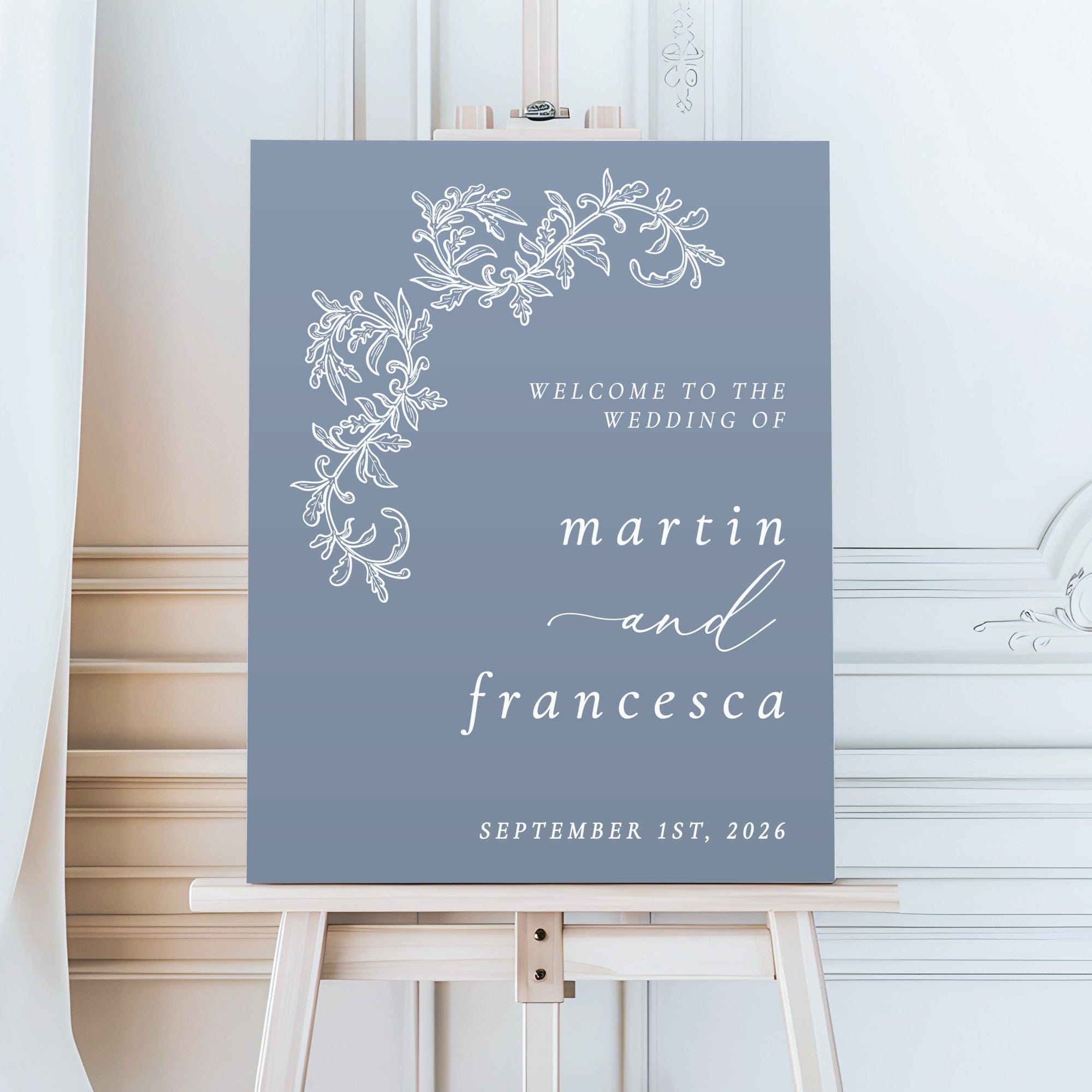 A rectangular welcome sign made from a rigid material in a blue-grey color, featuring elegant white lettering and a decorative floral vine design in the corner, intended to greet guests at a wedding event.