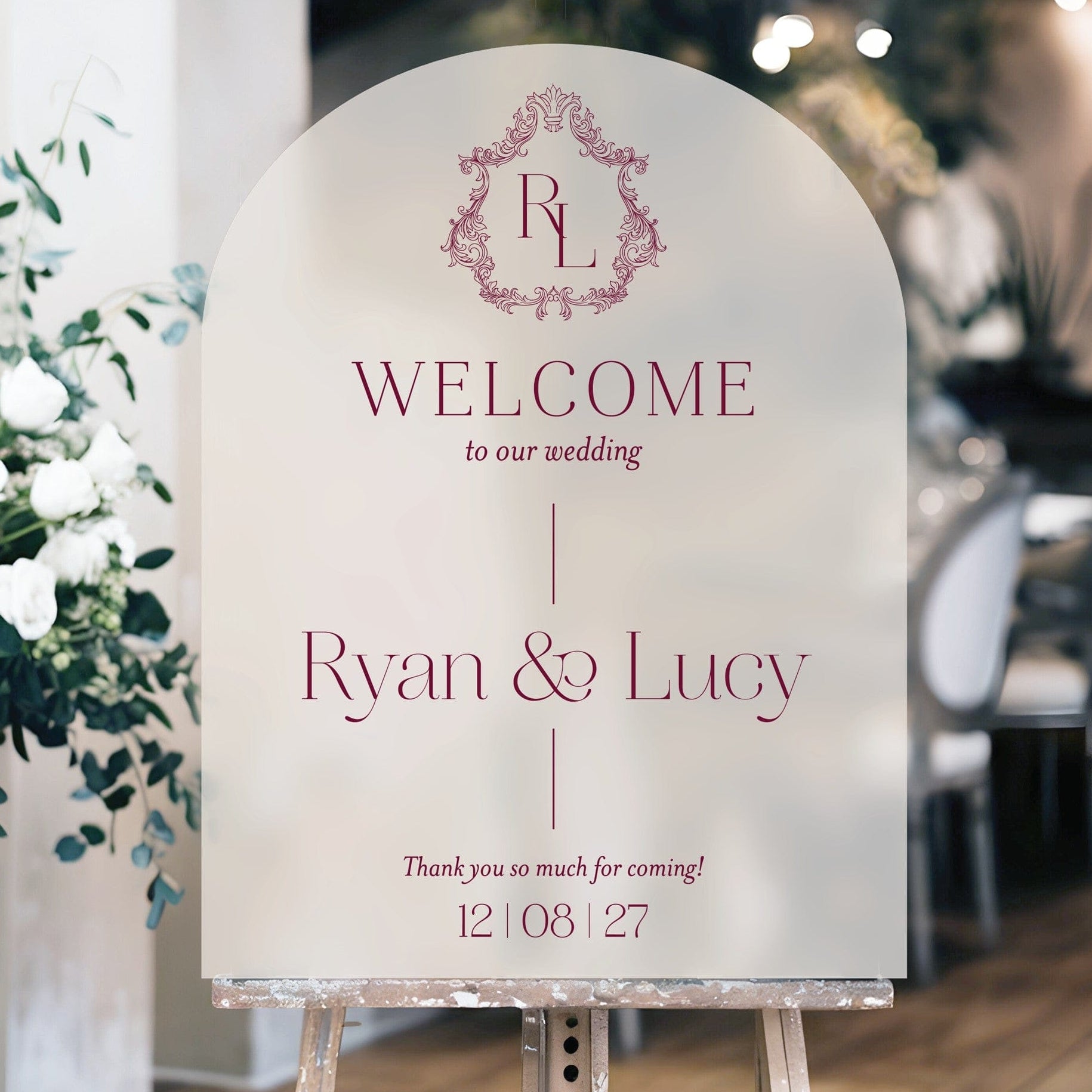 An elegant arched frosted wedding welcome sign featuring a decorative crest and personalized text in a deep maroon color, displayed on a wooden easel.