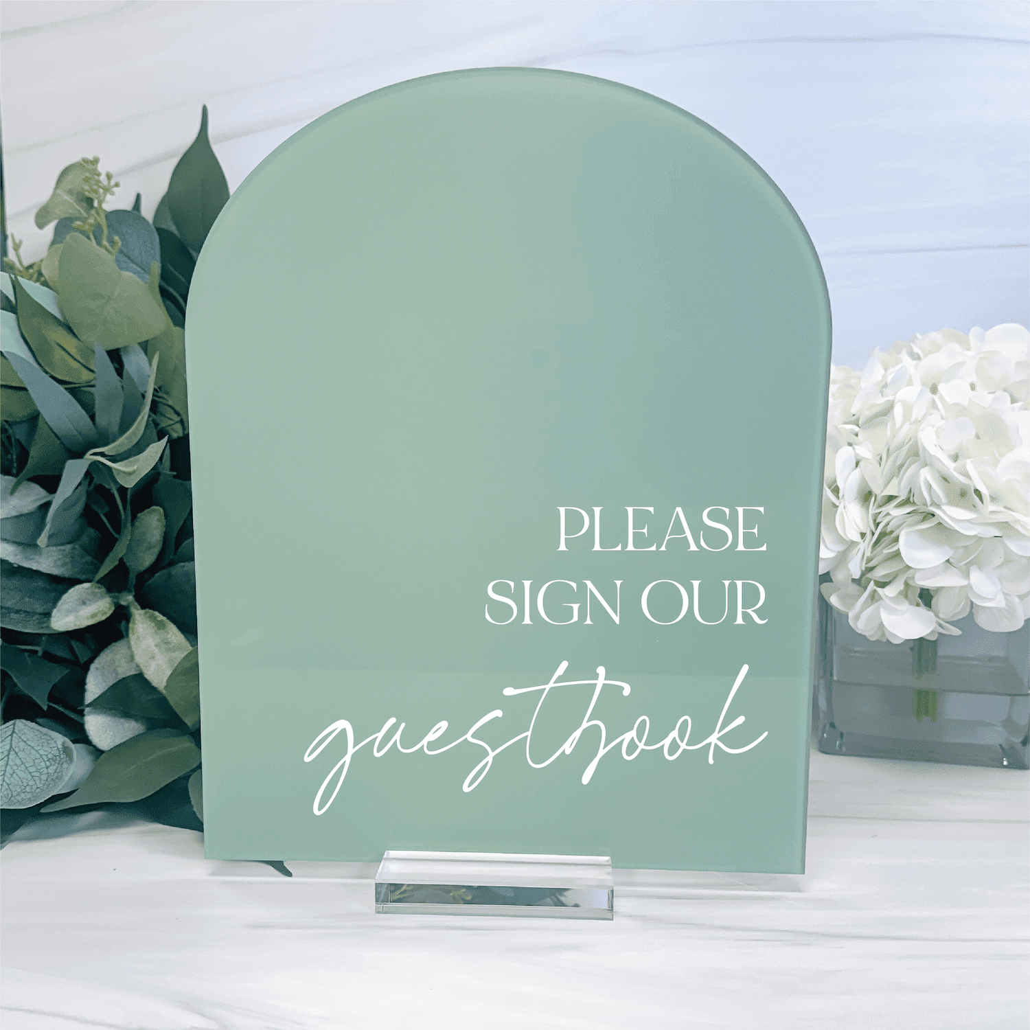 Sage green arched acrylic sign with white text reading "Please sign our guestbook" displayed with flowers and a pen holder.