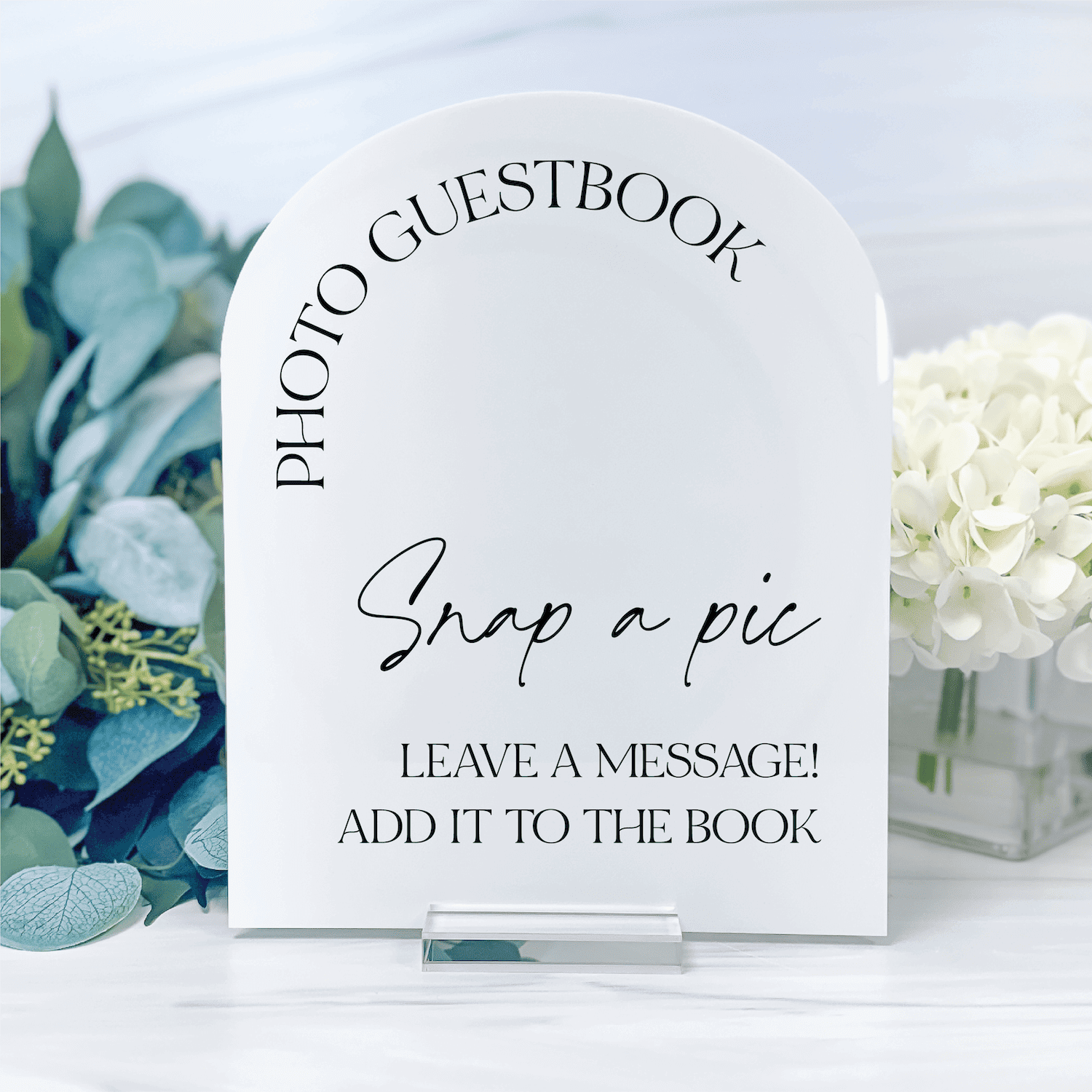White acrylic arch sign with "Photo Guestbook" at top, "Snap a pic" in script, and "Leave a message! Add it to the book" text below, displayed with greenery and white flowers.