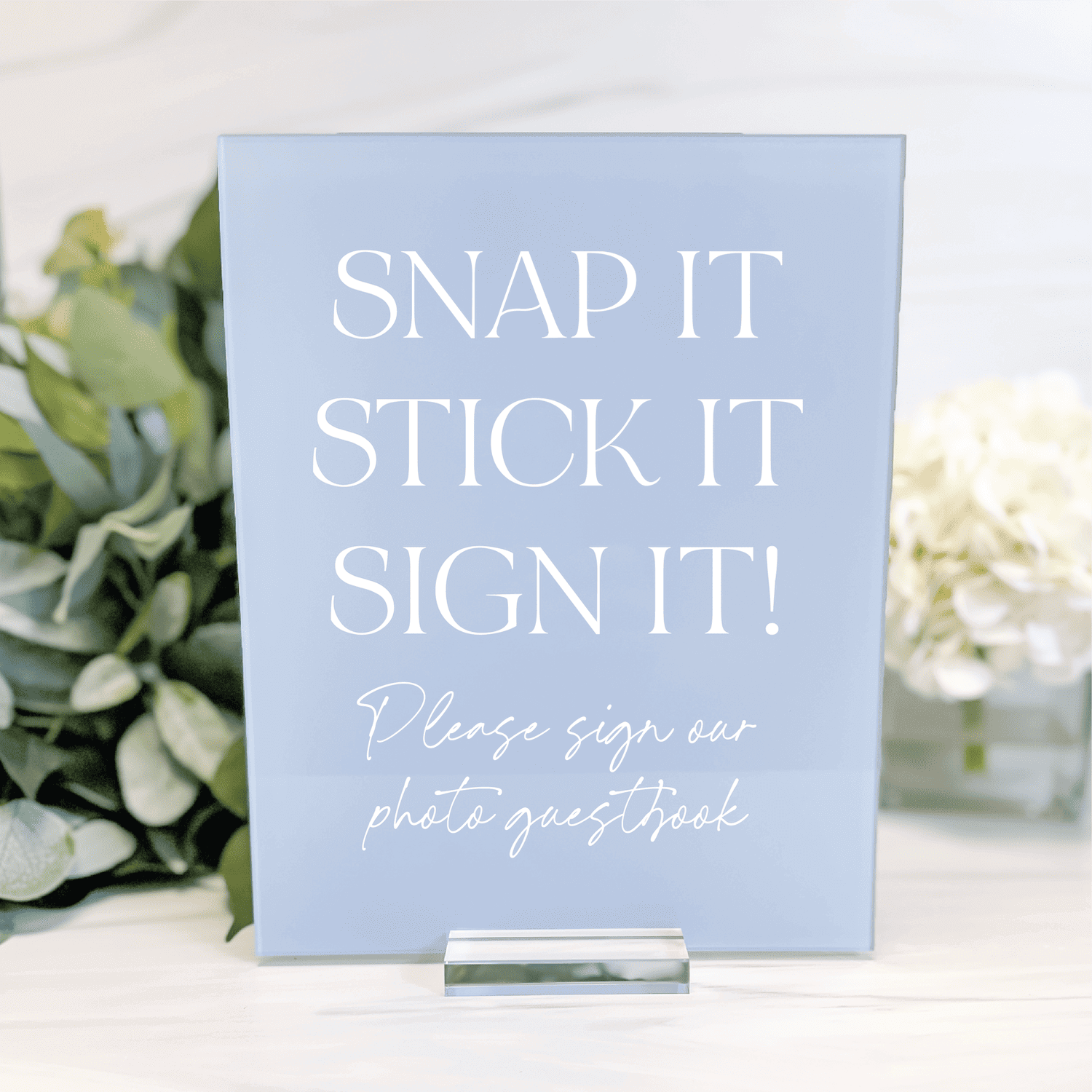 Light blue acrylic sign with white text reading "Snap it Stick it Sign it" and "Please sign our photo guestbook" displayed on white table with flowers.