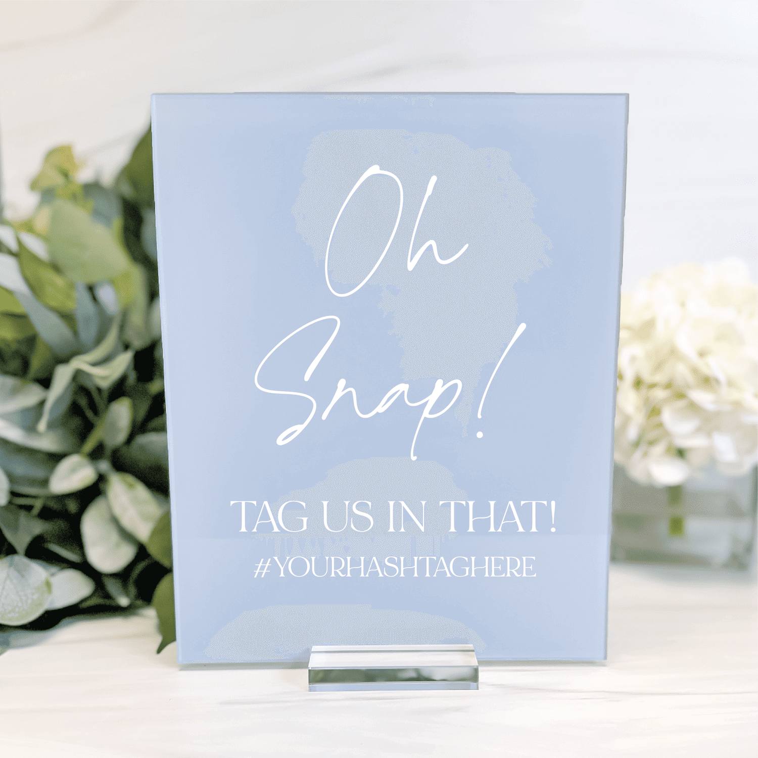 Light blue acrylic sign with white script text reading "Oh Snap! TAG US IN THAT! #YOURHASHTAGHERE" displayed on easel with greenery.