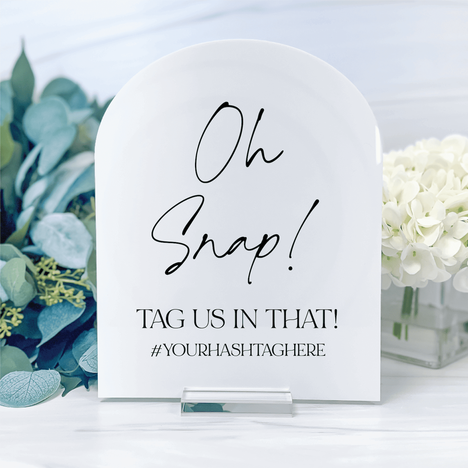 White acrylic arch sign with "Oh Snap!" text and "Tag us in that! #yourhashtaghere" message, displayed with eucalyptus and white flowers.