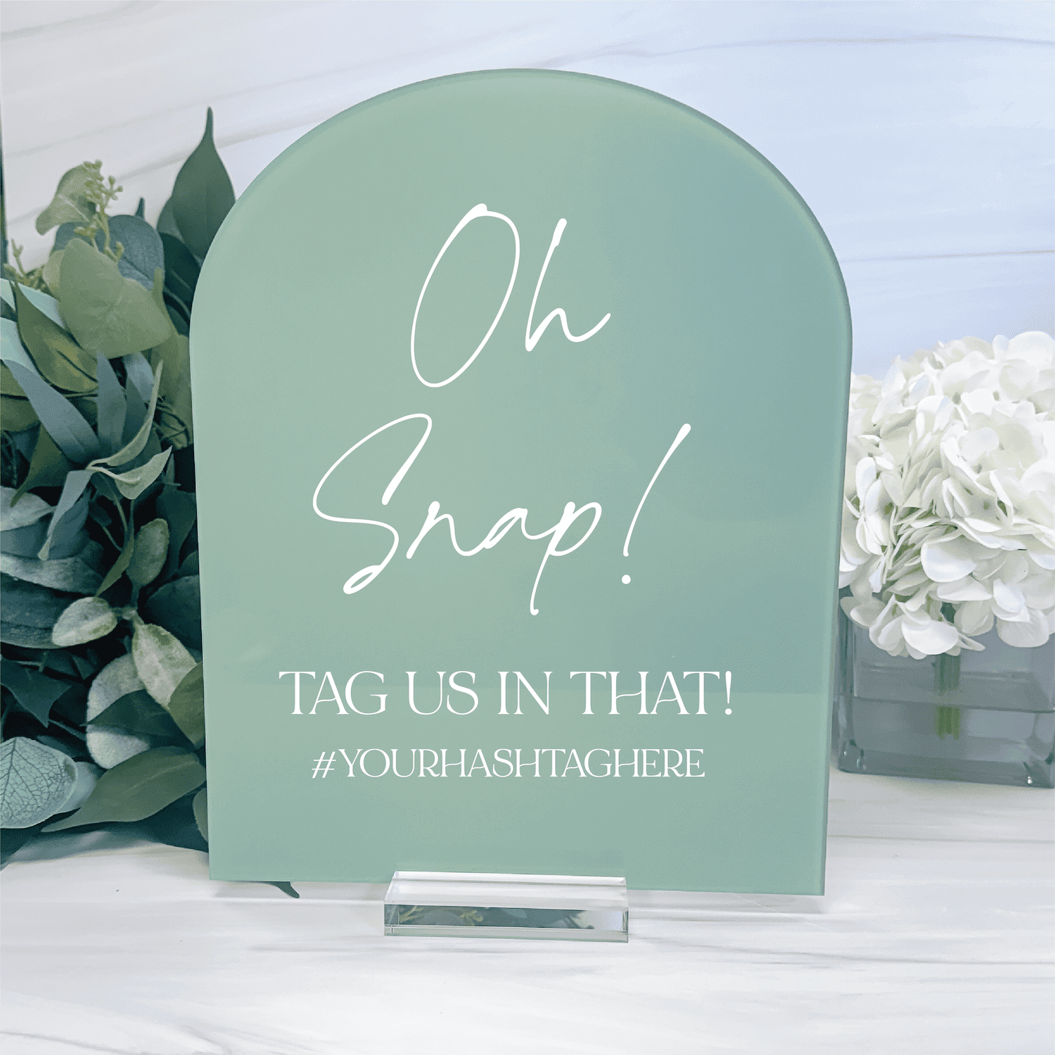 Sage green arch-shaped acrylic sign with white script text reading "Oh Snap! TAG US IN THAT! #YOURHASHTAGHERE" displayed with eucalyptus and white flowers.
