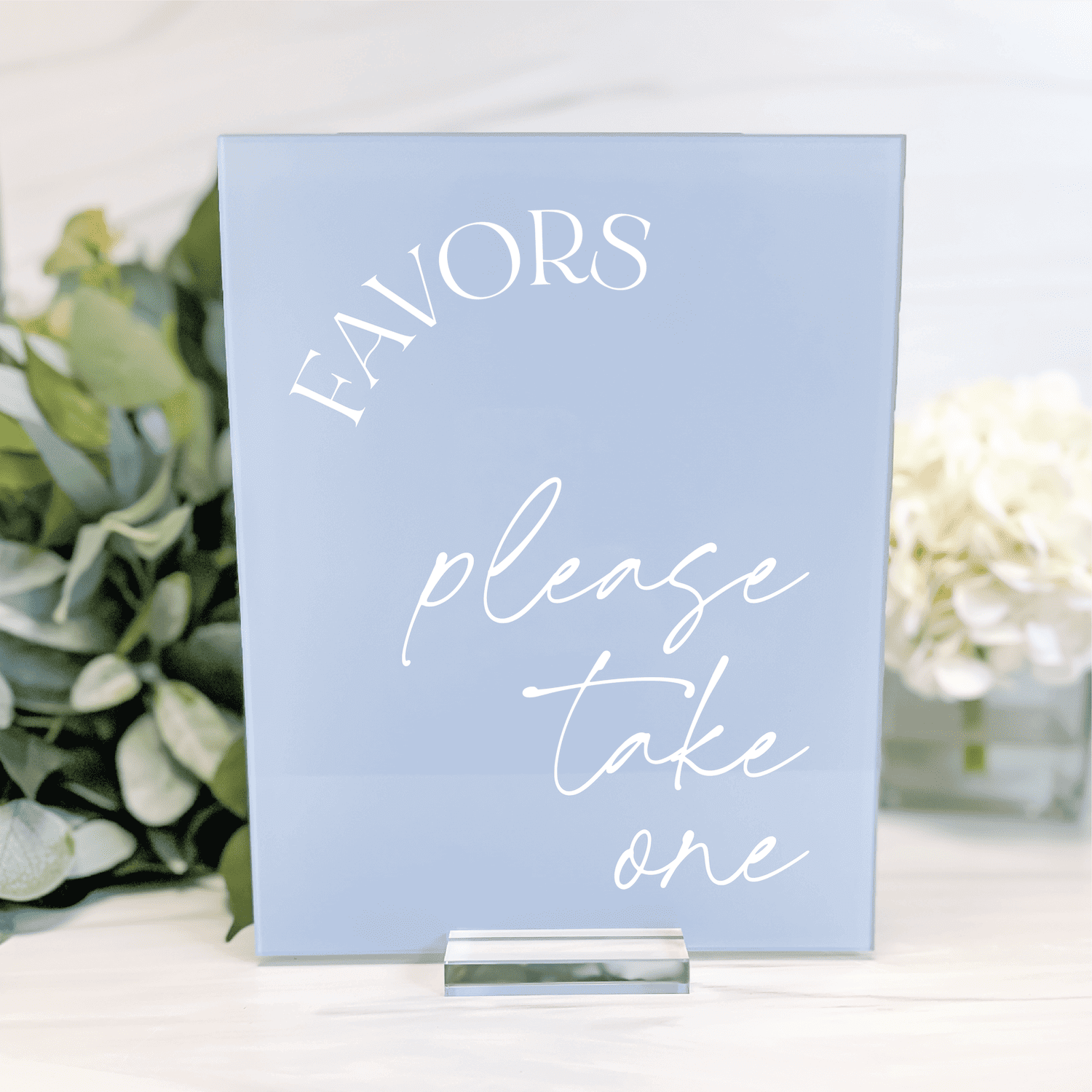 Light blue acrylic table sign with white text reading "FAVORS please take one" displayed on white surface with flowers.