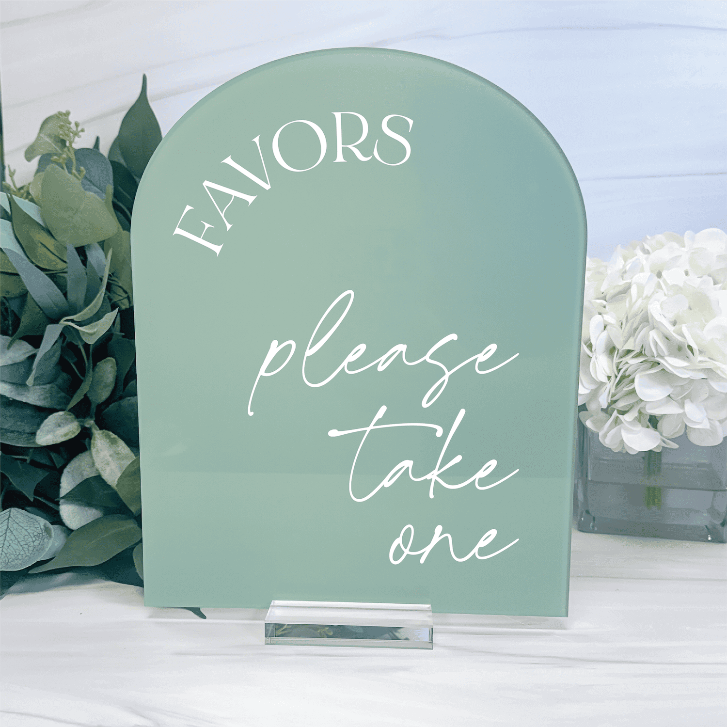 Sage green arched acrylic sign with white text reading "FAVORS please take one" displayed with floral arrangements.