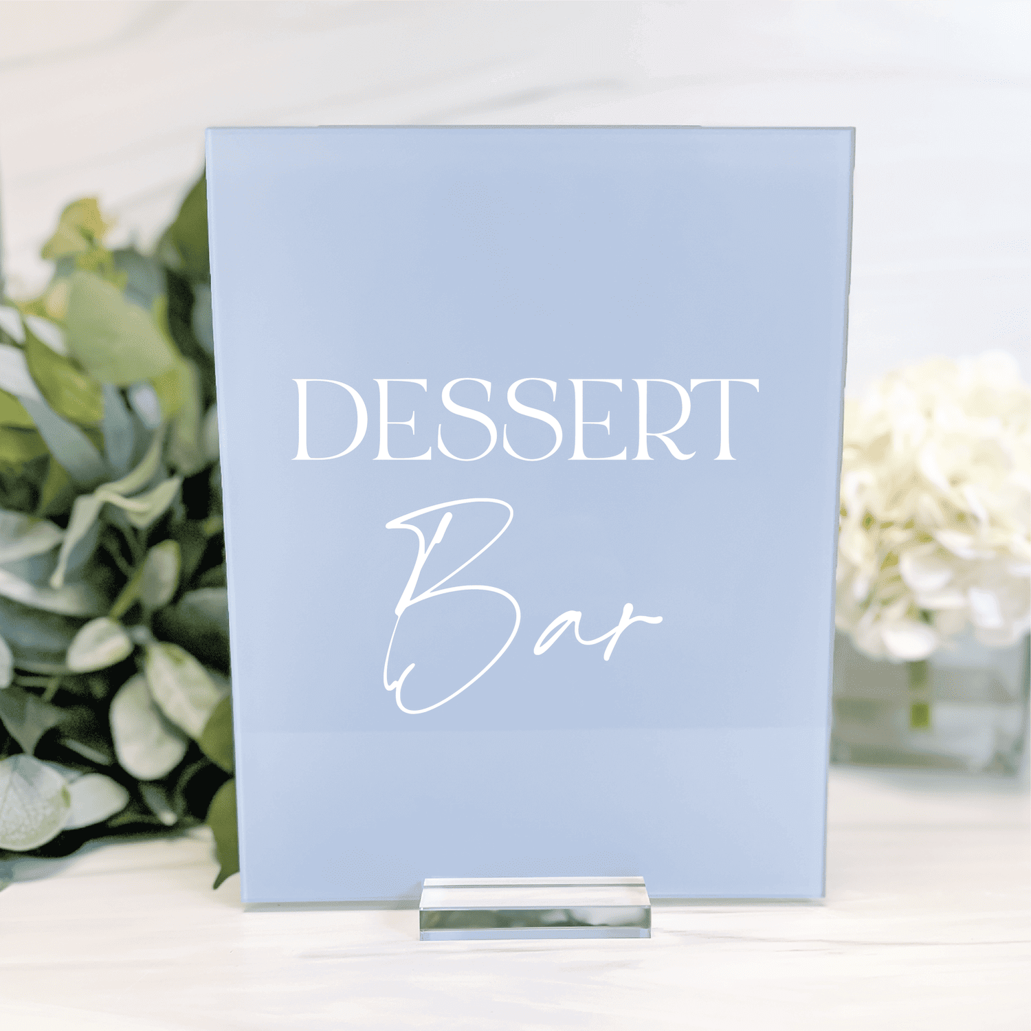 Light blue acrylic table sign with white text reading "DESSERT Bar" displayed on white surface with greenery and flowers.